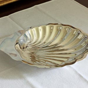 May include: A polished silver-toned metal dish shaped like a scallop shell. The dish features a series of radiating, curved ridges that extend from the base to the scalloped edge, creating a reflective, smooth surface with a metallic finish.