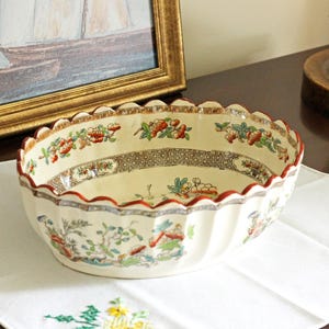 May include: An oval ceramic bowl with a cream-colored base, featuring a scalloped rim with a thin red edge. The exterior and interior are decorated with a repeating pattern of colorful floral motifs, green foliage, and brown geometric bands.