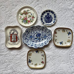 May include: A collection of six ceramic trinket dishes featuring various designs: a floral round dish, a blue and white Canada crest dish, a rectangular dish with the text La Tour d'Argent 1582, a scalloped blue bird dish, and two square floral dishes.