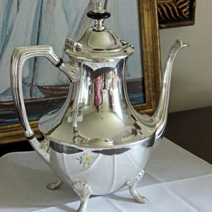 May include: A polished silver-plated teapot featuring a curved handle, a long spout, and a lid with a black finial. The body has a fluted design with a small floral decal near the base, supported by three ornate feet with shell-like detailing.