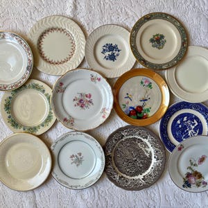 May include: A collection of twelve round ceramic plates featuring various designs, including floral patterns, blue and white willow-style prints, gold-rimmed edges, and solid cream surfaces, arranged together in a flat, overhead display.