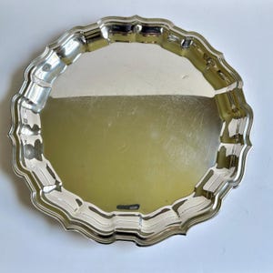 May include: A circular, silver-plated serving tray with a decorative, scalloped edge. The tray has a highly reflective surface and a wavy border. The design is simple and elegant, suitable for serving food or displaying items.