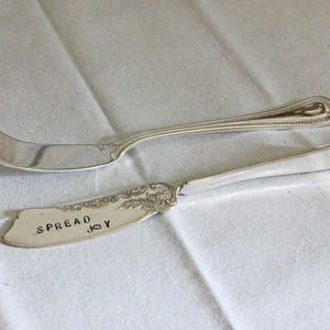 May include: Two silver-toned metal serving utensils. The top piece is a cheese knife with a three-pronged tip and a ribbed handle. The bottom piece is a flat spreader with an ornate floral pattern at the base and the text SPREAD JOY stamped on it.