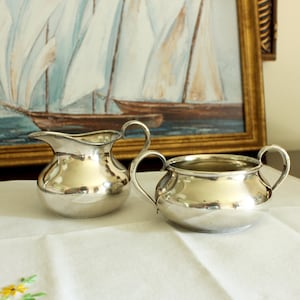 May include: A silver creamer and sugar bowl set. The creamer has a curved handle and a spout. The sugar bowl has two handles. Both pieces are highly polished and reflect the light. The set is placed on a white tablecloth in front of a framed painting.