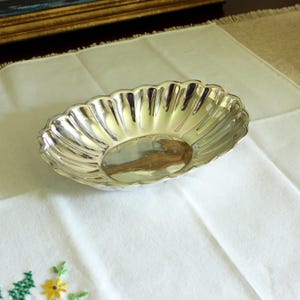 Puede incluir: Una fuente ovalada plateada con borde festoneado. La fuente es muy reflectante, con reflejos de luz visibles en su superficie. Se encuentra sobre un mantel blanco con detalles florales bordados, lo que sugiere un propósito decorativo o de servicio.
