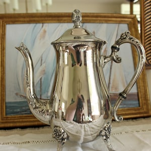May include: A polished silver-toned metal teapot featuring an ornate handle with black accents, a curved spout with floral detailing, a matching lid with a finial, and three decorative feet with flower motifs at the base of the body.