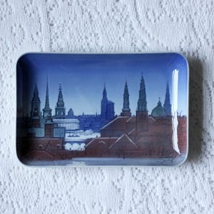 May include: A rectangular porcelain tray featuring a detailed illustration of a city skyline at twilight. The scene depicts brown-roofed buildings and various church spires set against a deep blue sky, with a smooth, glossy finish and rounded edges.
