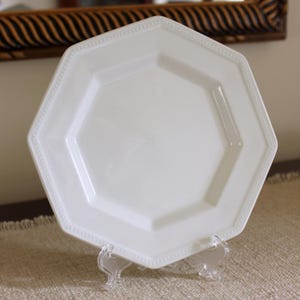 Puede incluir: Un plato blanco octogonal con un borde con cuentas. El plato se exhibe sobre un soporte de plástico transparente. El plato tiene un acabado brillante y está vacío. El fondo incluye un espejo decorativo.