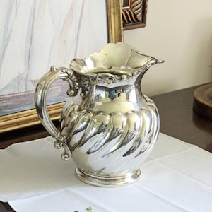 May include: A polished silver-tone pitcher featuring a rounded body with a swirling fluted pattern, a wide pouring spout, and an ornate curved handle. The metal surface is reflective, showing highlights and shadows across its curved form.