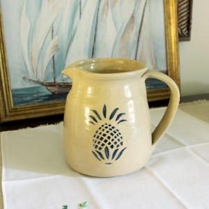 May include: A beige ceramic pitcher with a blue pineapple design. The pitcher has a handle and a spout for pouring. The background includes a framed painting of sailboats and a white cloth.