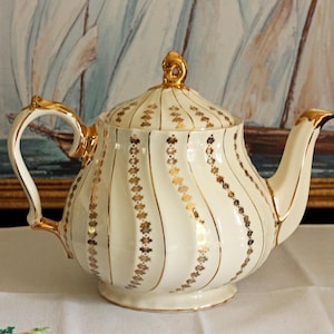 May include: A cream-colored porcelain teapot featuring a swirling ribbed body decorated with vertical gold floral patterns. The teapot includes a matching lid with a gold finial, a curved handle, and a spout, all accented with gold trim.