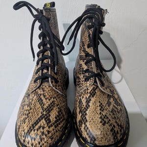 May include: A pair of brown and black snake-print leather boots with black laces. The boots have a classic lace-up design, a rounded toe, and a black sole with yellow stitching.