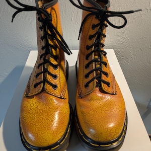 May include: A pair of orange and yellow floral-patterned boots with black laces. The boots have a glossy finish and a black sole. The top of the boots have the text "AirWair" in yellow.