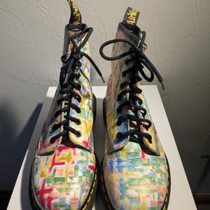 May include: A pair of colorful, patterned boots with black laces. The boots feature a vibrant design of overlapping rectangles in shades of red, yellow, blue, and green. The soles are black with yellow stitching.