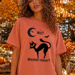 May include: A terracotta-colored short-sleeve t-shirt featuring a black graphic of a crescent moon, flying bats, and an arched cat with yellow eyes. The text reads "Moi?" above the cat and "Mischief-maker?" below the cat in black lettering.