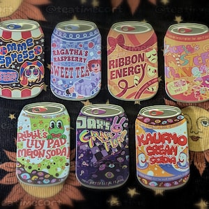 Puede incluir: Una colección de siete pines de esmalte rectangulares con forma de latas de refresco, cada uno con diseños gráficos coloridos únicos, ilustraciones de personajes y texto estilizado como Pomme Espresso, Ragatha's Raspberry Sweet Tea y Jax's Grape Float.
