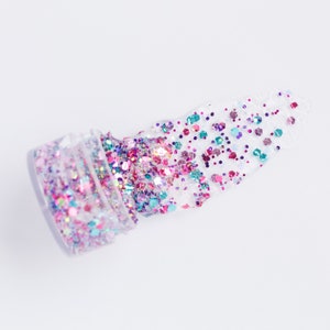May include: A clear plastic jar of colorful glitter is tipped over, spilling onto a white surface. The glitter is a mix of pink, purple, teal, and iridescent colors, with various shapes and sizes. The glitter is scattered in a thick, textured line.