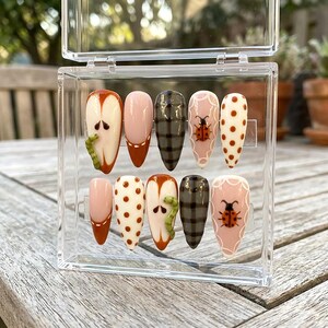 May include: A set of ten artificial nails in a clear plastic case. The nails feature various designs, including apple slices with green worms, pink and brown polka dots, plaid patterns, and ladybugs. The nails are in a variety of shapes and colors.