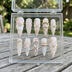 May include: A set of ten press-on nails in a clear plastic case labeled Silver14 nail. The nails are almond-shaped with a cream base, featuring 3D floral reliefs, gold metallic accents, iridescent shell fragments, and small white pearl beads.