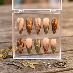May include: A set of ten almond-shaped press-on nails in a clear plastic case. The nails feature a color palette of nude, bronze, and metallic gold with intricate raised leaf patterns, starburst accents, and small embedded circular stones.