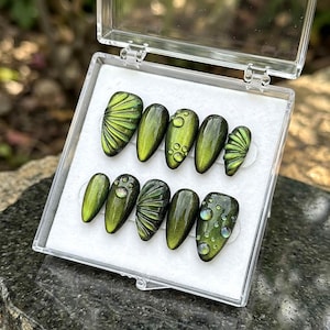 May include: A set of ten green press-on nails in a clear plastic case. The nails feature various designs, including fan-like patterns and water droplet accents. The nails have an oval shape and a glossy finish.