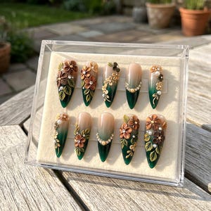 May include: A set of ten artificial nails in a clear display case. Each nail features a gradient design, transitioning from a dark green base to a nude or light pink tip. The nails are adorned with 3D floral and pearl embellishments in shades of brown, gold, and green.