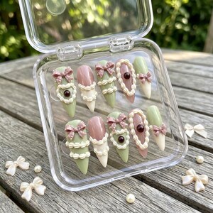 May include: A set of ten press-on nails in a clear plastic case, featuring a color palette of sage green, dusty rose, and cream. The nails are decorated with three-dimensional white piped icing details, small brown beads, and tiny polka-dot bows.