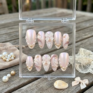 May include: A set of ten almond-shaped press-on nails with a pearlescent finish, displayed in a clear plastic case. The nails feature 3D white bows, intricate white line art, and small cream-colored pearls arranged in various decorative patterns.