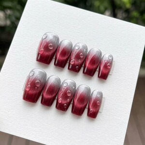 May include: Set of ten press-on nails with a gradient design. The nails transition from a silver color at the tips to a deep red at the base. Each nail features small, clear, droplet-shaped embellishments.