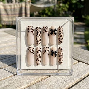 May include: A set of ten artificial nails in a clear display case. The nails feature a nude base with leopard print accents and black bow details. The nails are arranged in two rows of five.