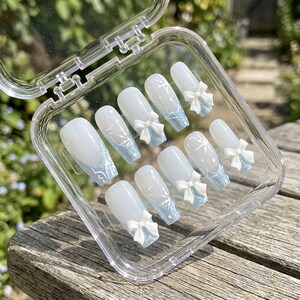 May include: A set of ten press-on nails in a clear plastic storage case. Each nail features a milky white base with a light blue glittery French tip, white snowflake patterns, and a small, three-dimensional white bow attached to the surface.
