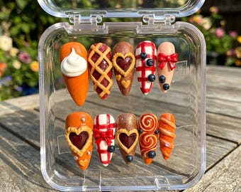 May include: A set of ten artificial nails in a clear plastic case. The nails feature various designs, including a whipped cream swirl, lattice pie crust, heart-shaped filling, red plaid, and bows. Some nails have small blueberry accents.