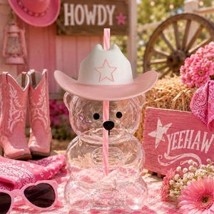 May include: A clear, bear-shaped plastic tumbler with a white cowboy hat lid featuring a pink star outline. A pink and white striped straw extends through the center of the hat. The bear has black circular eyes and a black nose.