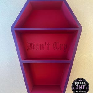 Puede incluir: Un estante de pared con forma de ataúd con un marco exterior morado y un interior rosa intenso. El estante cuenta con dos compartimentos horizontales, con el texto "Don't Cry" grabado en una fuente gótica en el panel posterior de la sección superior.