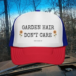 May include: A red, white, and blue trucker hat. The hat has a white front panel with the text "GARDEN HAIR DON'T CARE" in black, sunflower graphics, a red brim, and a blue mesh back. The text "DIRT ROAD AF" is also visible.