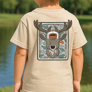 May include: A tan t-shirt featuring a cartoon deer wearing a hat, set against a camouflage background. The deer has large antlers and a smiling face. The design is framed in a green border.