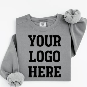 May include: A grey crewneck sweatshirt featuring the black text "YOUR LOGO HERE" printed in a bold, block font on the center chest. The garment has long sleeves with ribbed cuffs and a Comfort Colors brand label visible at the inner neckline.