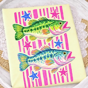 May include: A decorative item featuring two colorful fish illustrations. The fish are green and blue, with open mouths, set against a light yellow background with pink vertical stripes and blue and pink star accents. The artwork has a whimsical, hand-painted style.
