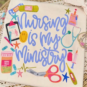 May include: Cream-colored t-shirt with the phrase "Nursing is my Ministry" in blue script. The shirt features colorful illustrations of medical supplies, including a stethoscope, chart, pills, and a syringe. The design includes stars and a smiley face.