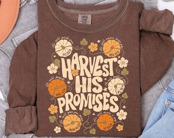 May include: A brown long-sleeve crewneck sweatshirt featuring a graphic print with the text "HARVEST HIS PROMISES" surrounded by illustrations of pumpkins, flowers, and vines, along with small script text including Bible verses.