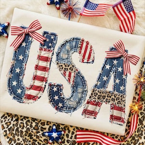 May include: Cream-colored shirt with the word "USA" appliqued in denim, red and white striped fabric, and leopard print. Each letter is embellished with a red, white, and blue striped bow. Patriotic theme.