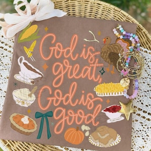 Könnte beinhalten: Ein braunes T-Shirt mit dem orangefarbenen Text God is great, God is good, umgeben von Illustrationen eines Truthahns, Mais, einer Soßenschüssel, eines Kuchens, eines Kürbisses und Kartoffelbrei, verziert mit einer hellrosa Schleife und einem Perlenarmband.
