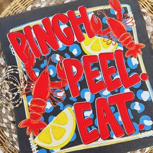 May include: Black t-shirt with a red and white design featuring the words "Pinch Peel Eat", red crawfish, and lemon slices. The background has a blue and white leopard print pattern. The design is framed with a gold border.