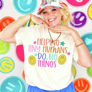 May include: A cream-colored short-sleeve t-shirt featuring the colorful text "HELPING TINY HUMANS DO BIG THINGS" in pink, orange, and blue. The design includes two yellow smiley faces and small star accents in pink, blue, and green.
