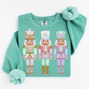 May include: A seafoam green crewneck sweatshirt featuring a graphic of three nutcrackers in pastel pink, mint, and lavender colors with gold glitter accents. The inner neck label displays the text Comfort Colors. The long sleeves are cuffed.