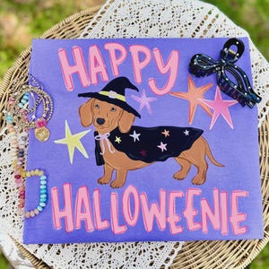 Puede incluir: Una camiseta morada con un perro salchicha marrón que lleva un sombrero de bruja negro y una capa negra con estrellas de colores. El texto HAPPY HALLOWEENIE aparece en letras rosas. La camiseta está decorada con estrellas rosas y amarillas.