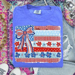 May include: A periwinkle blue Comfort Colors sweatshirt with a patriotic flag design. The flag design includes a bow, polka dots, gingham, and floral accents in red, white, and blue. The sweatshirt is folded on top of a pair of blue jeans.