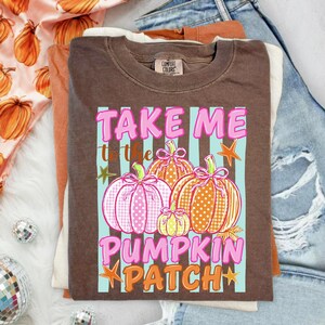 Könnte beinhalten: Ein braunes T-Shirt mit einem Aufdruck, der den Text TAKE ME TO THE PUMP KIN PATCH zeigt. Das Design umfasst drei Kürbisse in Rosa, Orange und Gelb mit Punktemustern und rosa Schleifen vor einem Hintergrund aus hellblauen Streifen.