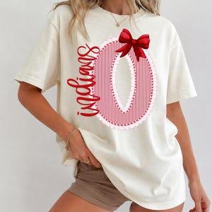 Puede incluir: Una camiseta de manga corta color crema con un gráfico grande de la letra O con rayas verticales rojas y un borde de encaje blanco. La palabra Indians está escrita en cursiva roja sobre la letra, rematada con un lazo rojo.