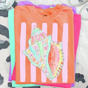 Könnte beinhalten: Ein orangefarbenes T-Shirt mit einem großen, farbenfrohen Muscheldesign und vertikalen rosa Streifen. Die Muschel ist mit verschiedenen Meereskreaturen verziert. Das T-Shirt ist mit anderen bunten Shirts gestapelt.